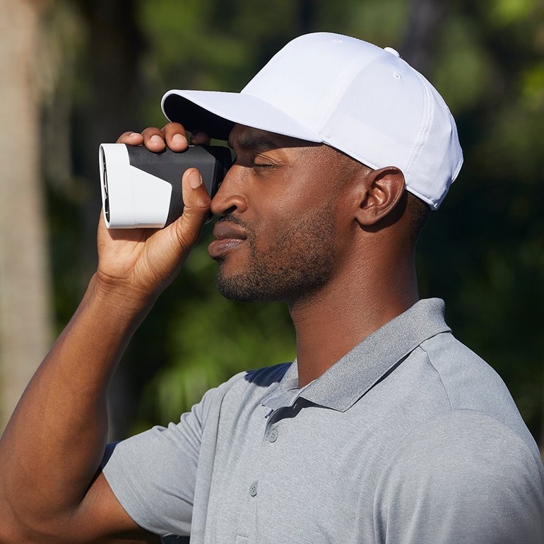 A golfer wearing a white cap and gray polo shirt is using a golf rangefinder to measure the distance on the golf course.