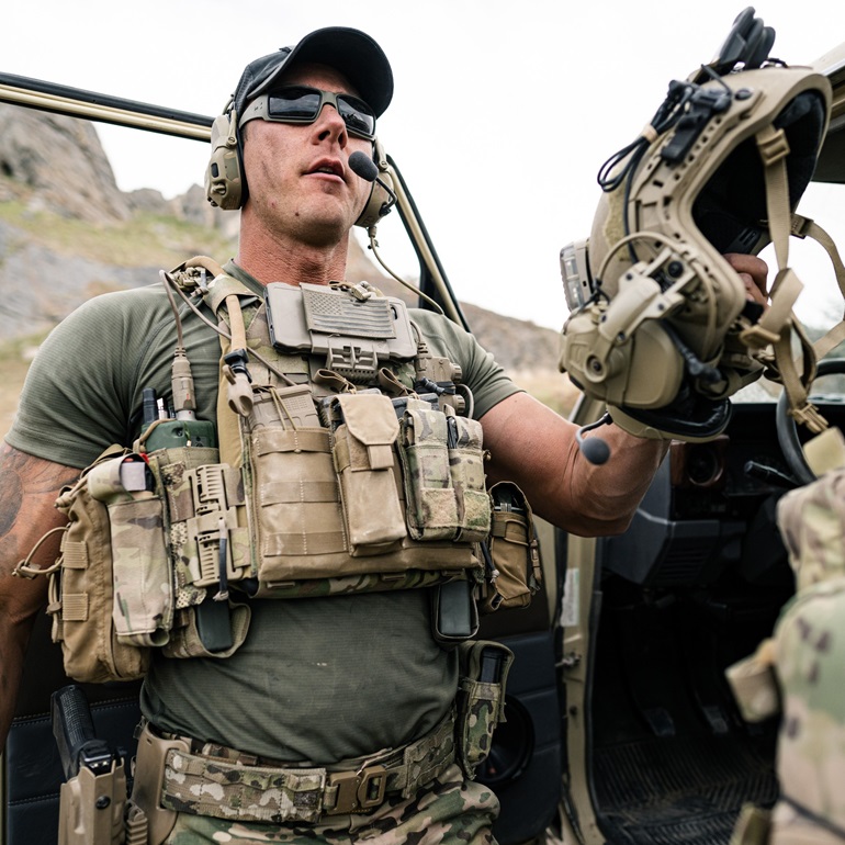 A soldier equipped with tactical gear, including a plate carrier with various pouches and a headset. He is holding a helmet in one hand, wearing sunglasses, and appears to be in a rugged outdoor setting, possibly preparing for a mission.