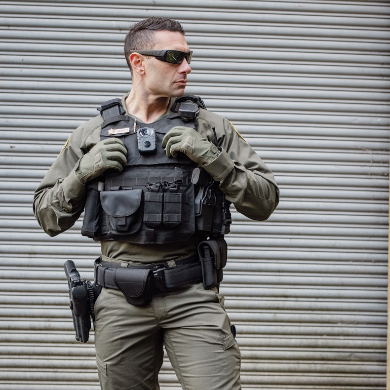 A tactical officer dressed in full gear, including a black plate carrier vest and green uniform. He is wearing sunglasses and gloves, standing in a ready stance in front of a corrugated metal background, likely preparing for an operation.