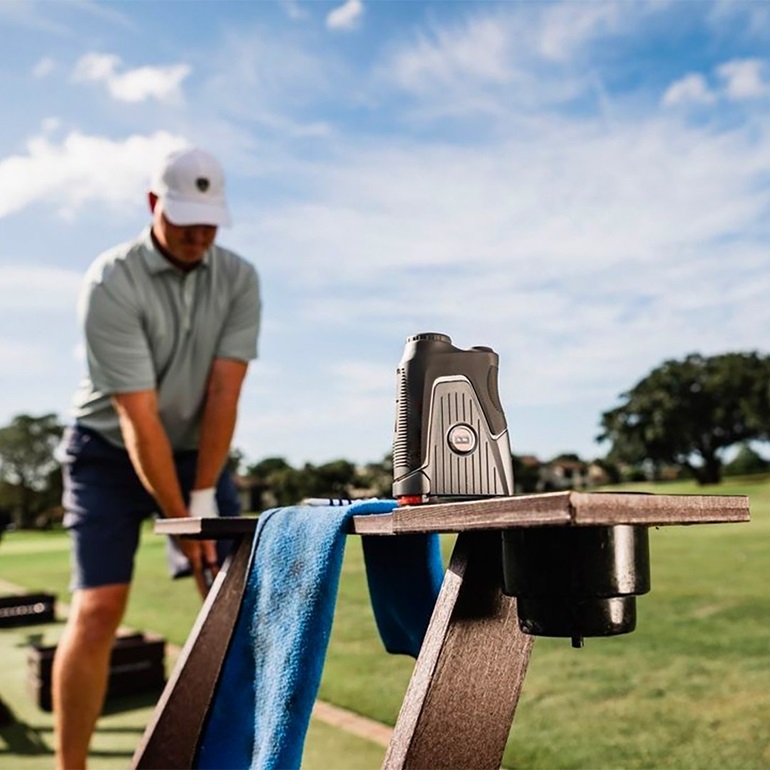 A golfer prepares to swing while a golf rangefinder rests on a bench nearby, ready for use on the sunny golf course.